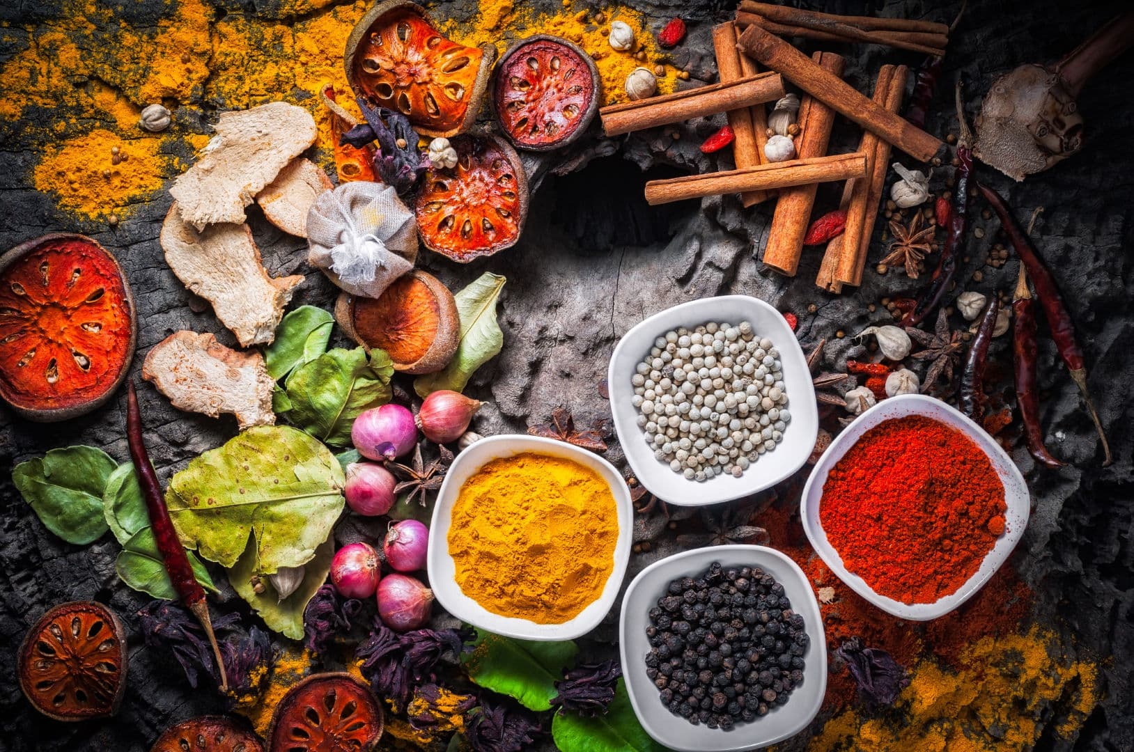 Bowls of turmeric, paprika and black and white peppercorns among cinnamon sticks, star anise and dried chillies